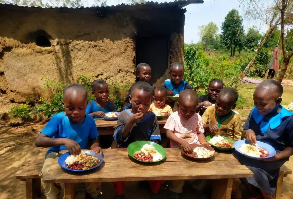  Mehrere Kinder sitzen im Freien an einem Holztisch und essen gemeinsam eine Mahlzeit. Im Hintergrund sind weitere Kinder und ein einfaches Lehmhaus zu sehen.